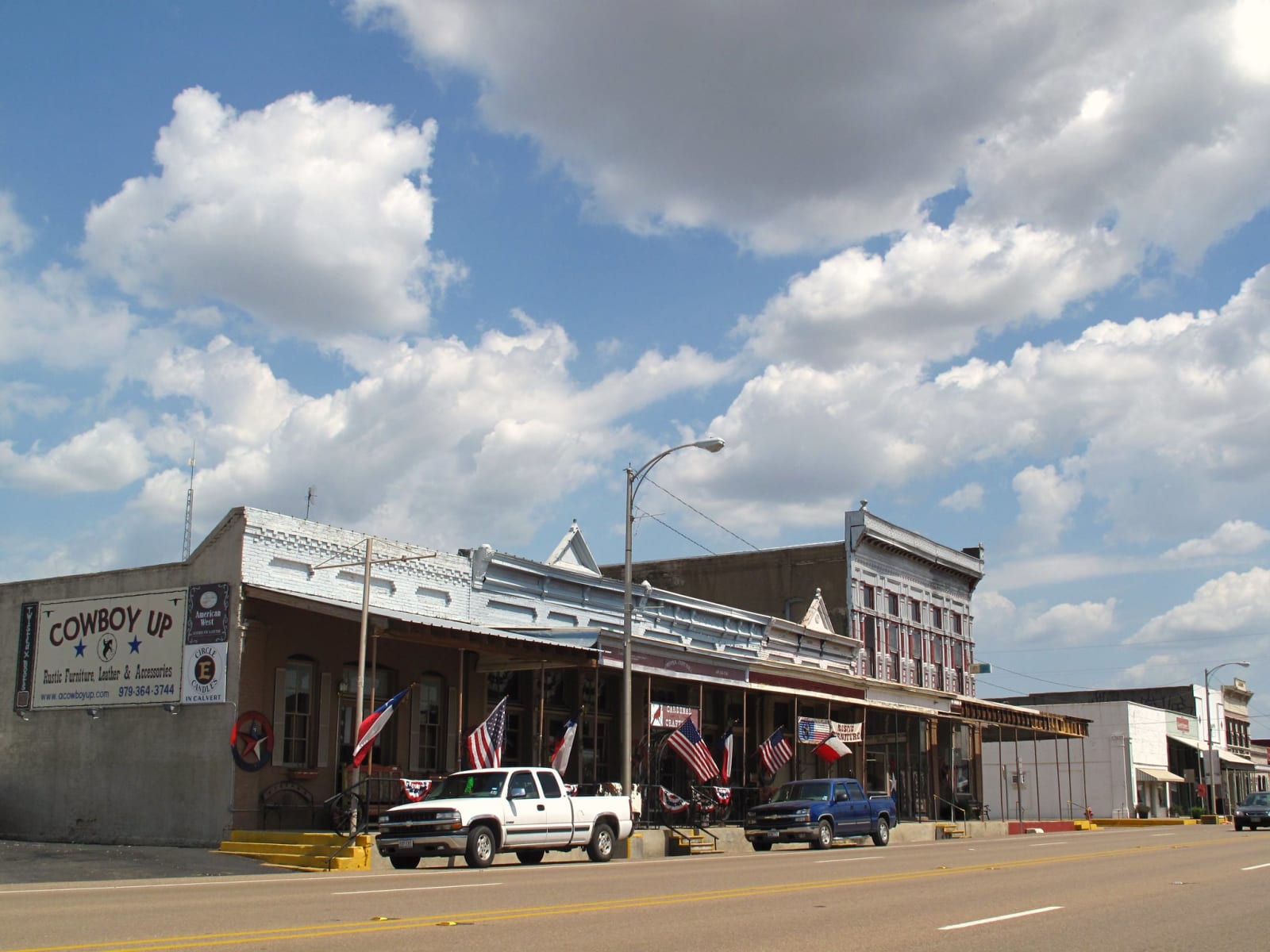 Main Street in Calvert, with brick storefronts and a two-story building with a long porch under a cloud-dotted sky.
