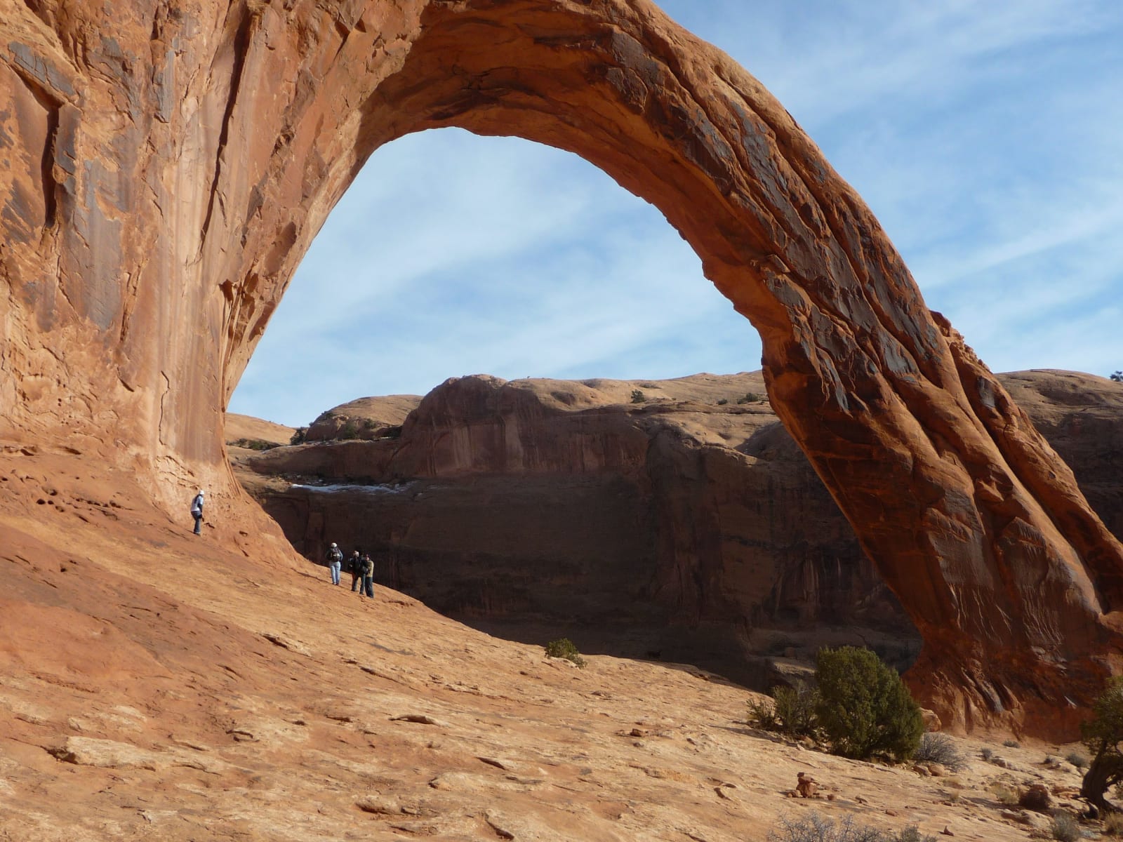 A massive sandstone arch springing from a cliff wall on the left and sweeping down to the right, deep blue sky showing through the opening. Four small figures stand on the bare slickrock slope below it, giving the scale, with a red rock bench behind.