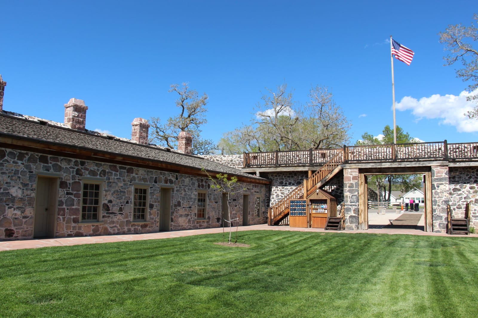 The courtyard inside the fort, looking across a mown lawn to a long single-story range built of dark volcanic stone with brick chimneys, green doors and a brick walk. A timber gallery runs above an open gateway on the right, and a flag flies from a pole beyond the wall.