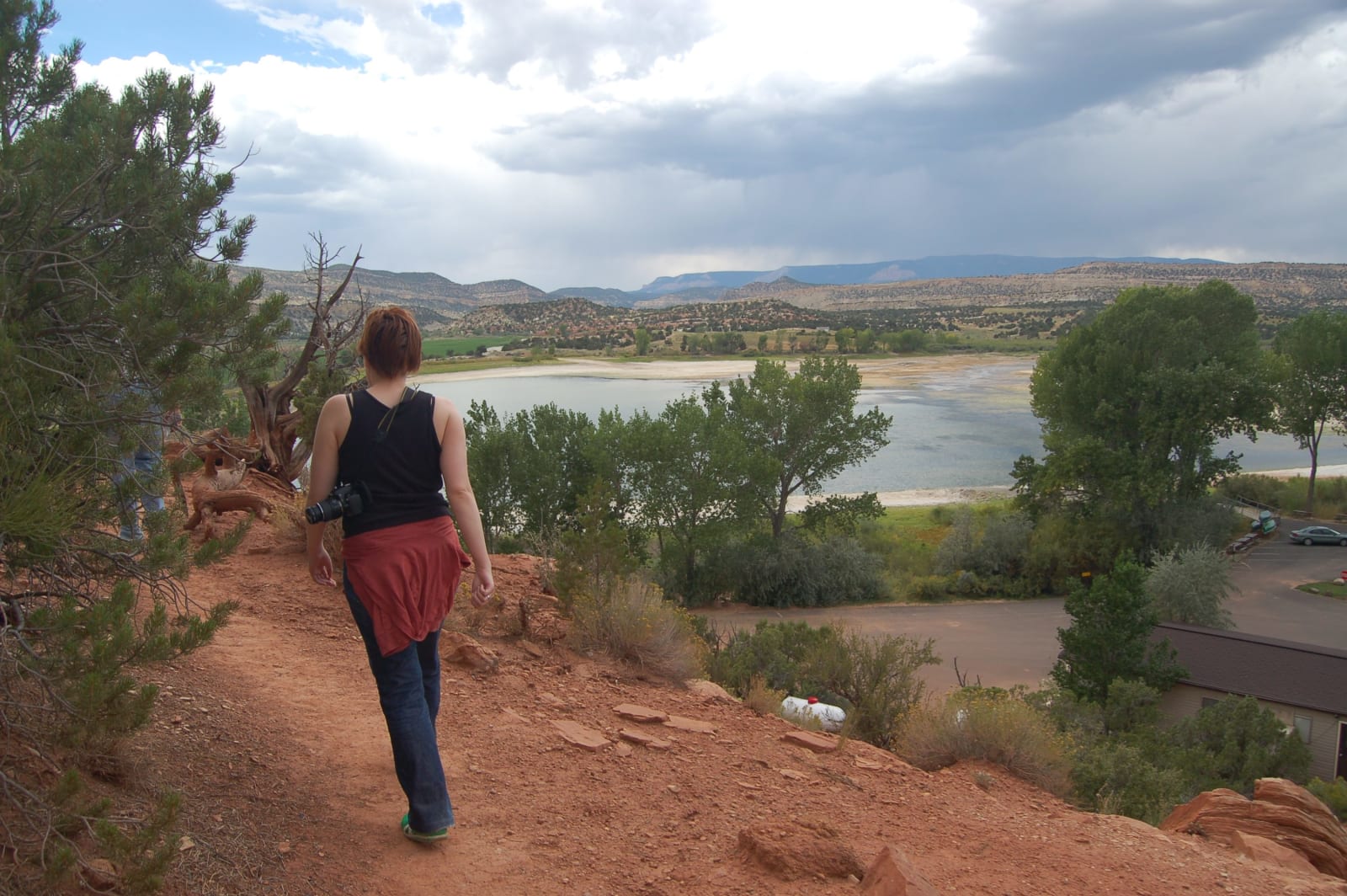 A hiker with a camera walking away down a red dirt trail toward Wide Hollow Reservoir, seen from the slope above — a juniper at left with a weathered log lying beside it, a second walker partly hidden behind the branches, and cottonwoods along the shoreline below. The reservoir's pale margin curves back to low benchland and mesas on the horizon under heavy grey cloud.
