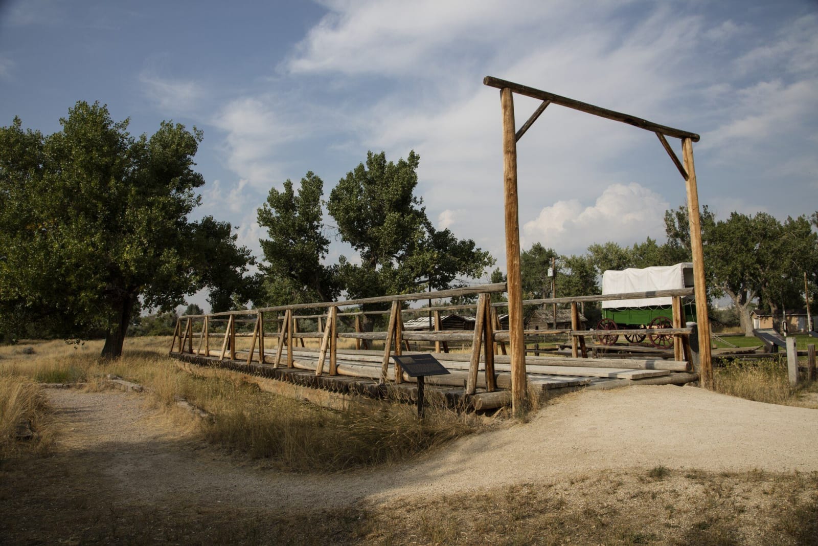 The reconstructed Guinard toll bridge at Fort Caspar, a timber deck on log cribs crossing a dry channel toward a covered wagon, cottonwoods behind