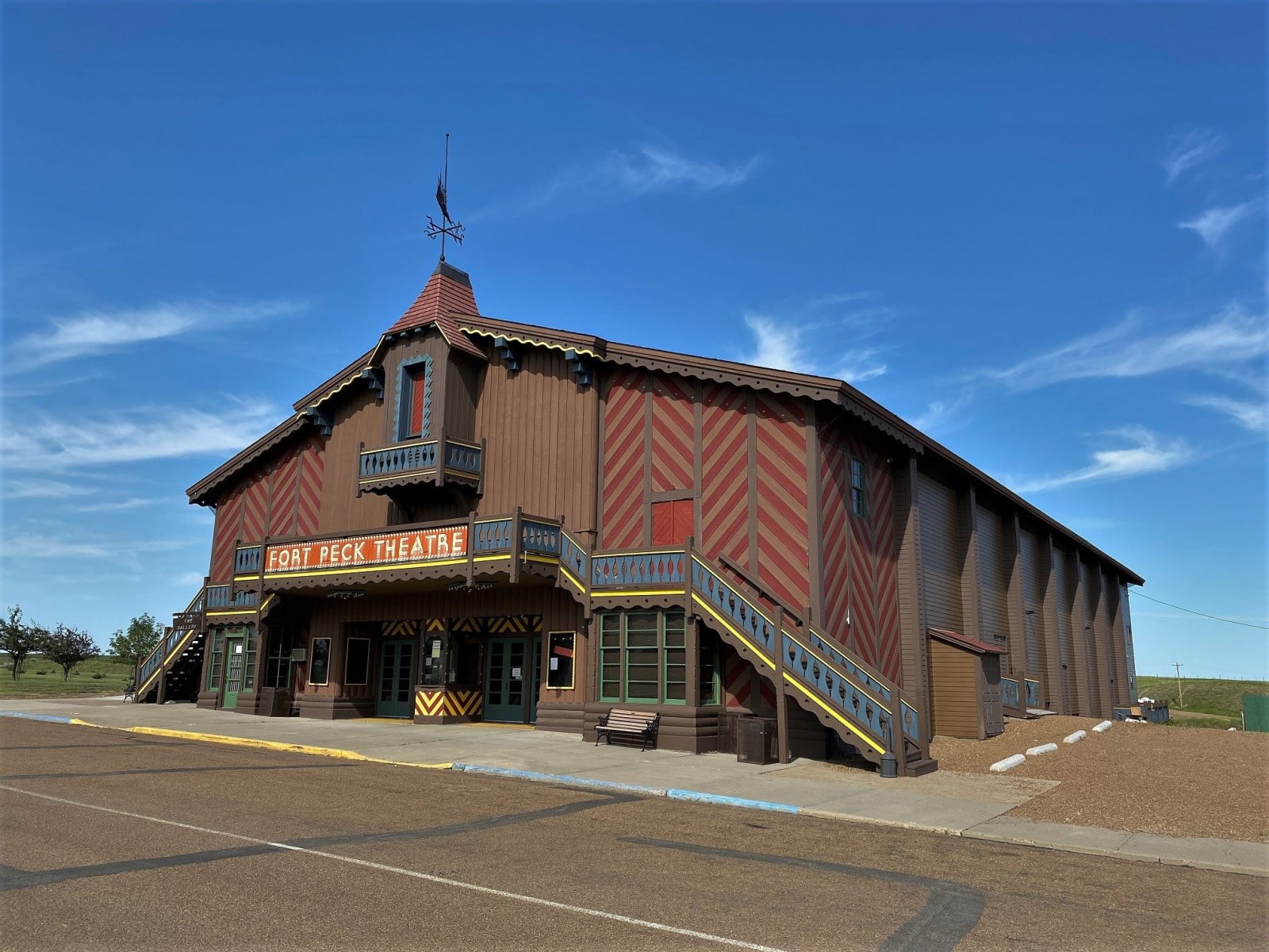 The Fort Peck Theatre, a big Swiss chalet-style hall of dark timber with red chevron panels and outside stairs, under a blue sky