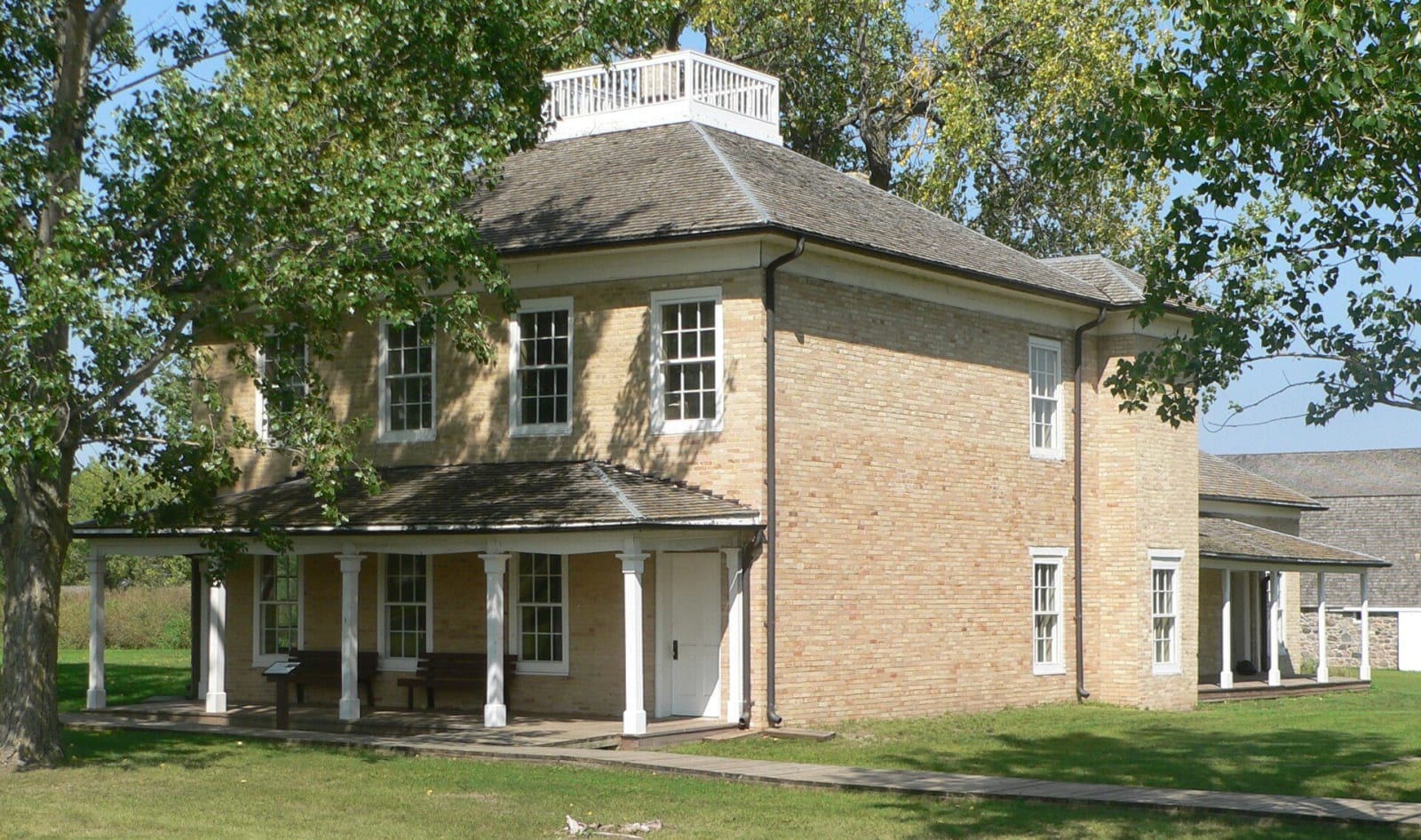 The two-story brick hospital at Fort Sisseton with its columned porch and rooftop lookout, under shade trees