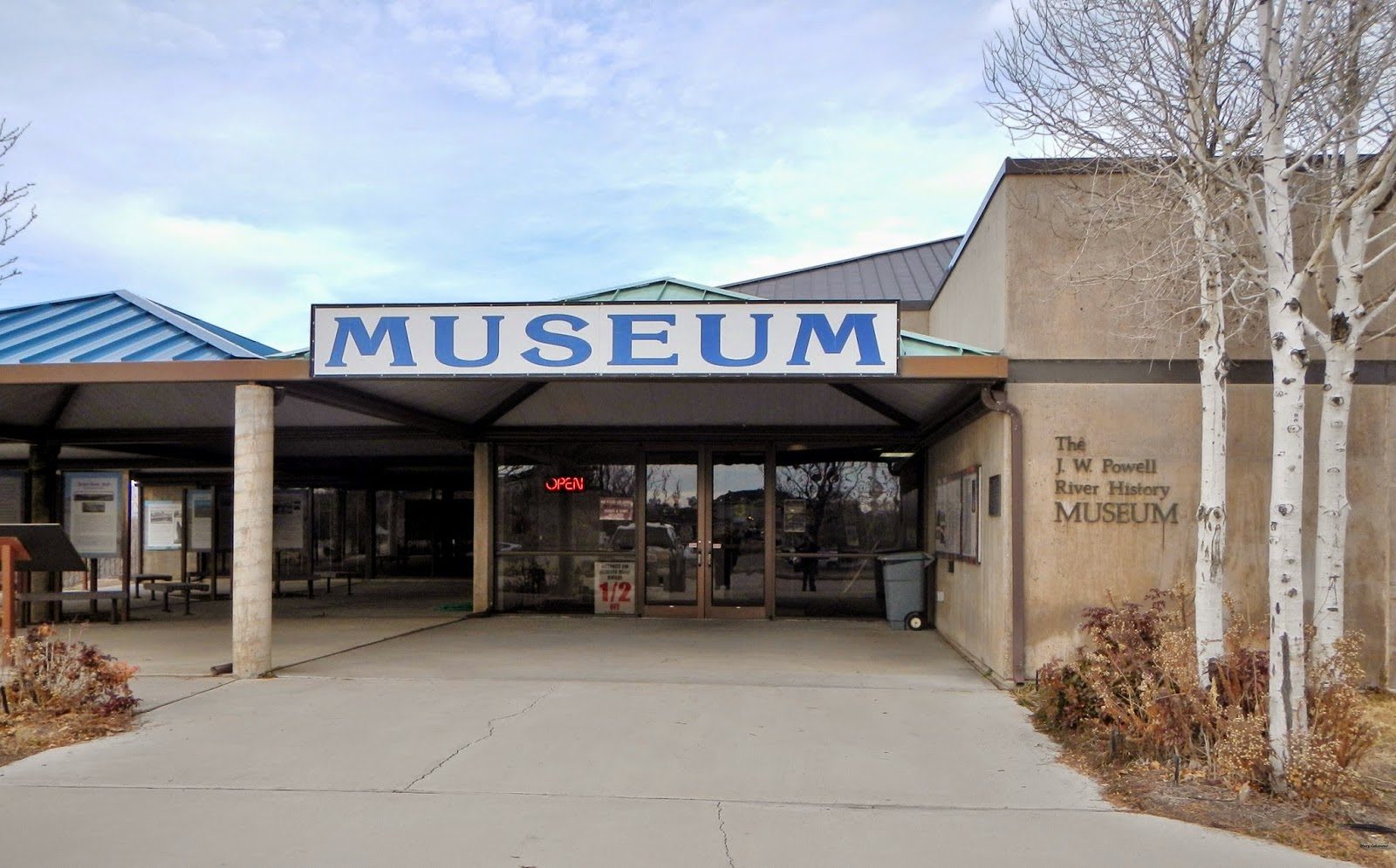 The entrance to the John Wesley Powell River History Museum in Green River — a canopy over glass doors carrying a large blue-on-white MUSEUM sign, with the museum's full name cut into the stucco wall at the right. Blue and green metal roofs rise behind, interpretive panels stand under the canopy at the left, and bare winter aspens screen the corner of the building.