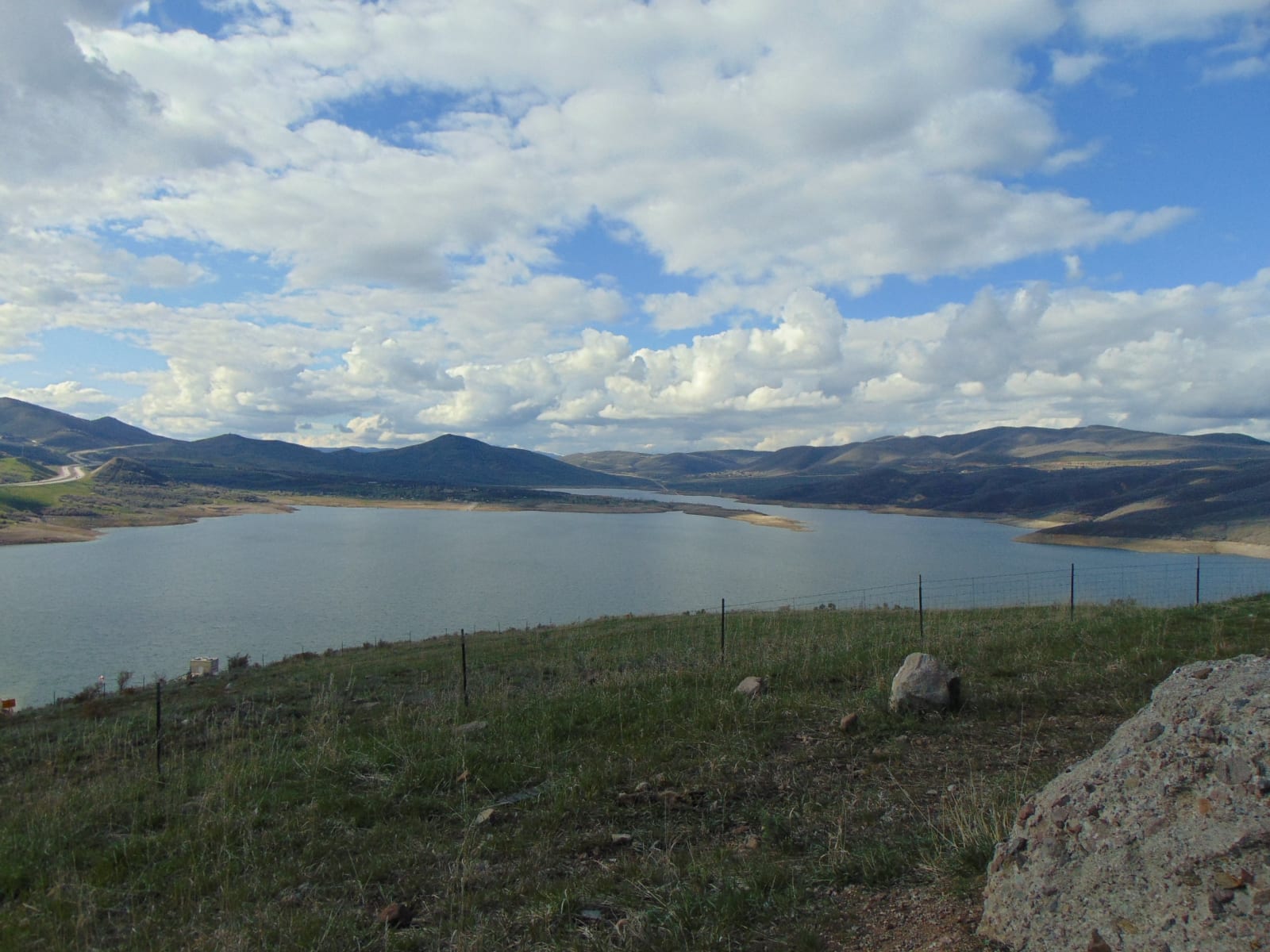 A reservoir filling a broad valley, its pale exposed shoreline showing well above the waterline, with low rounded hills running around it. A wire fence crosses a grassy bluff in the foreground and tall cumulus stands over the ridges.