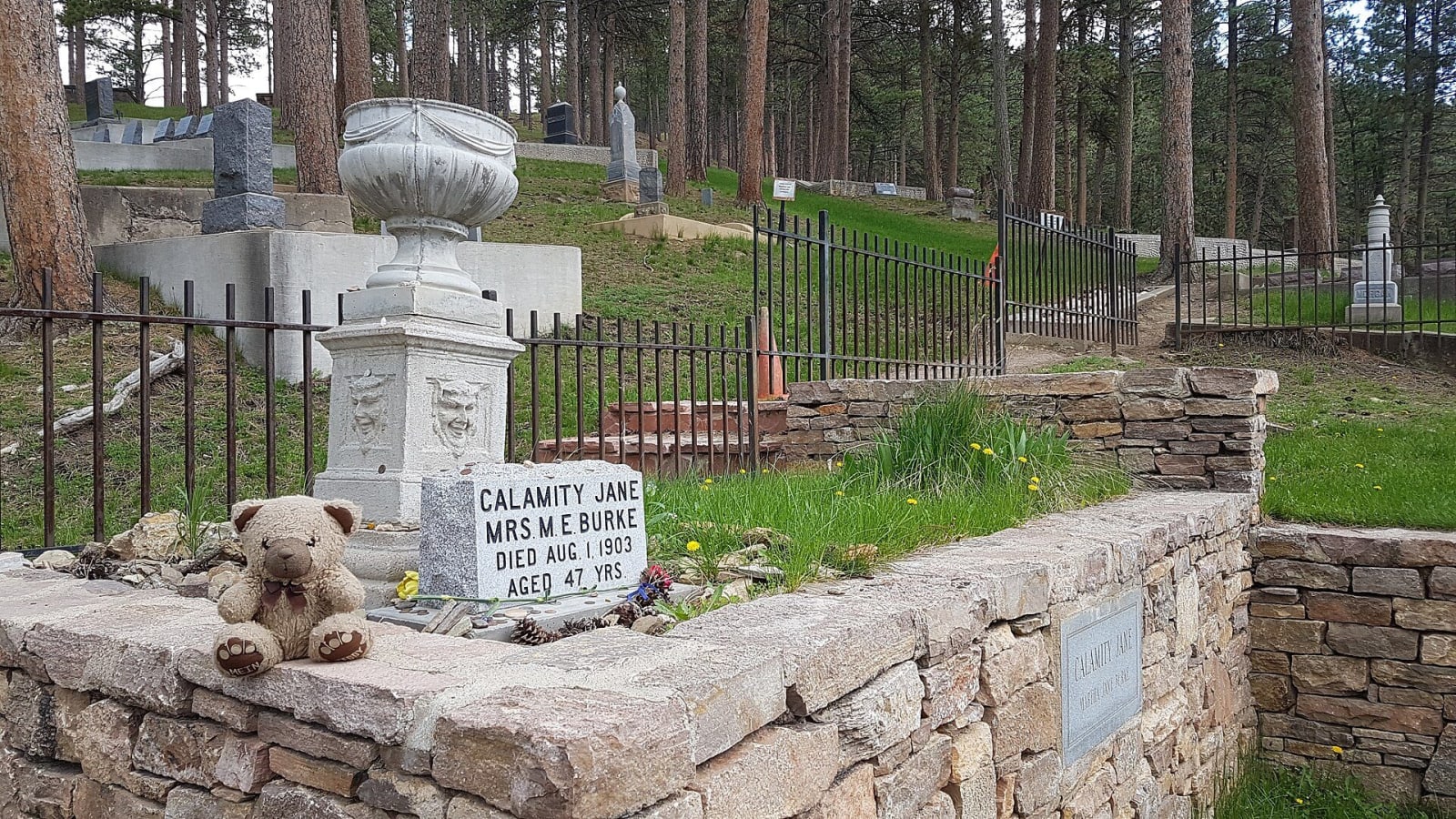 Calamity Jane's headstone behind a low stone wall in Mount Moriah Cemetery, with fenced plots and pines beyond