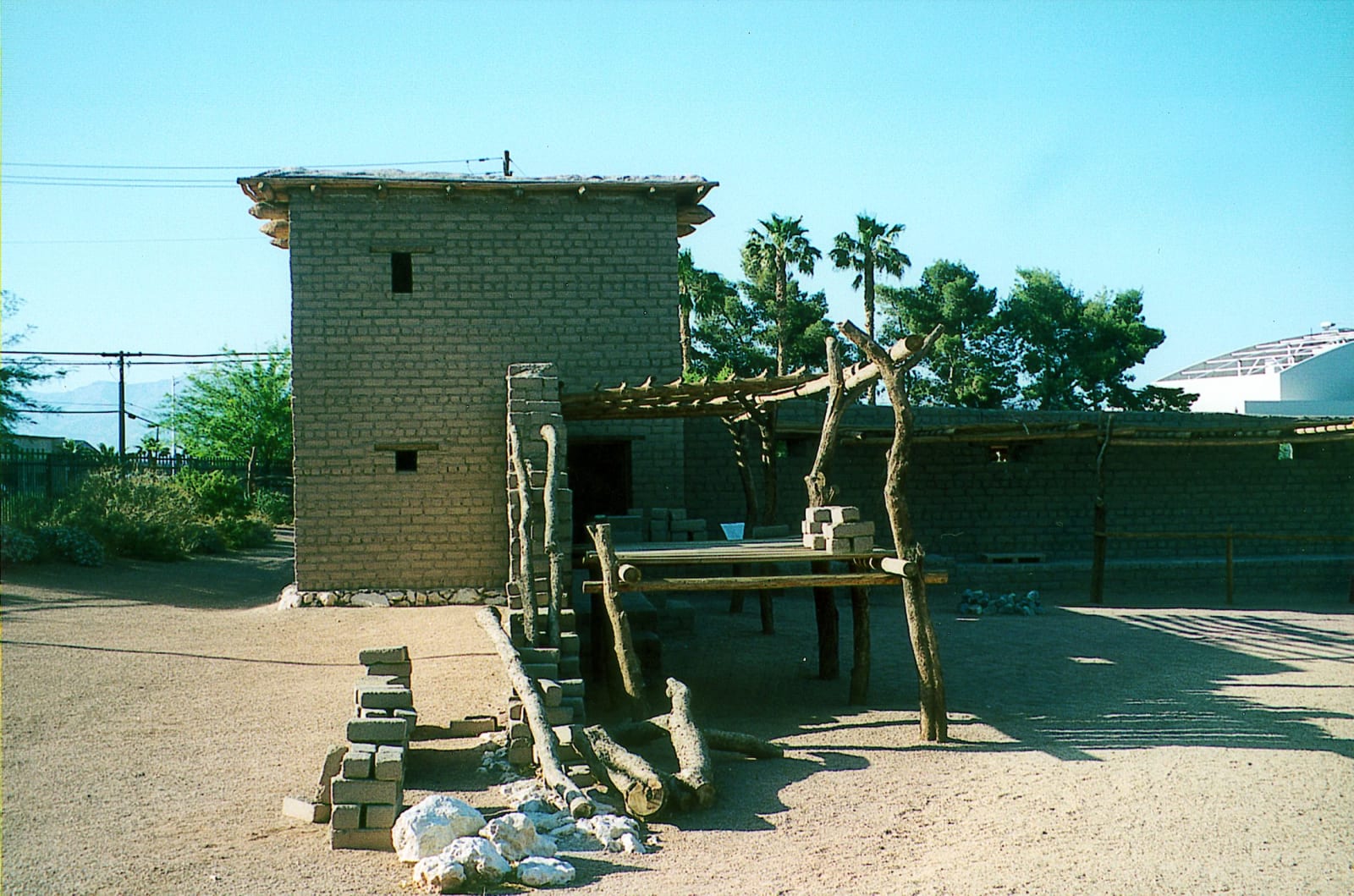 A two-storey adobe building with small square window openings at the Old Las Vegas Mormon Fort, a ramada of rough cottonwood posts and beams standing in front of it, courses of adobe bricks laid out to dry in the foreground and a modern white building visible over the wall behind.