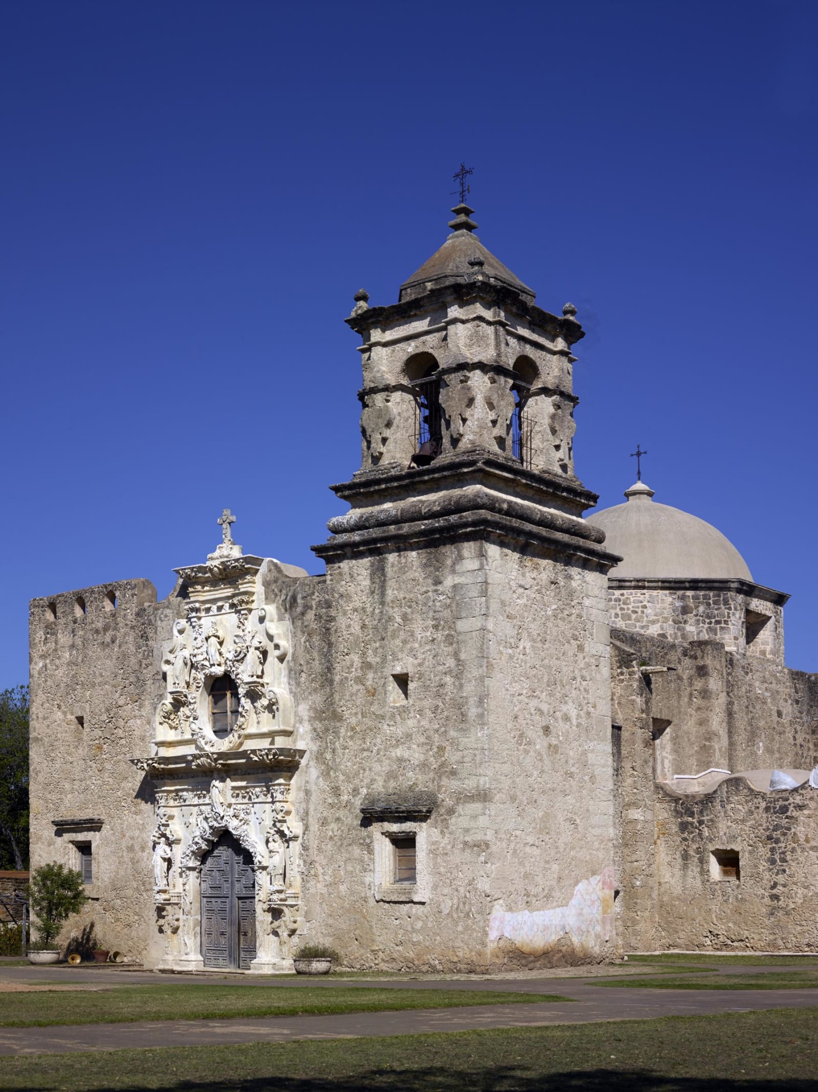 The stone bell tower, carved entrance and dome of the church at Mission San Jose in San Antonio under a clear sky.