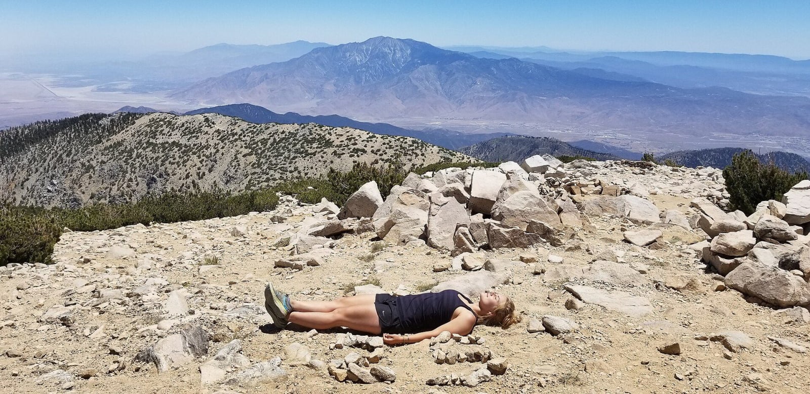 The bare rock summit of San Gorgonio Mountain above the tree line, a hiker resting among the stones, San Jacinto Peak across the pass beyond.