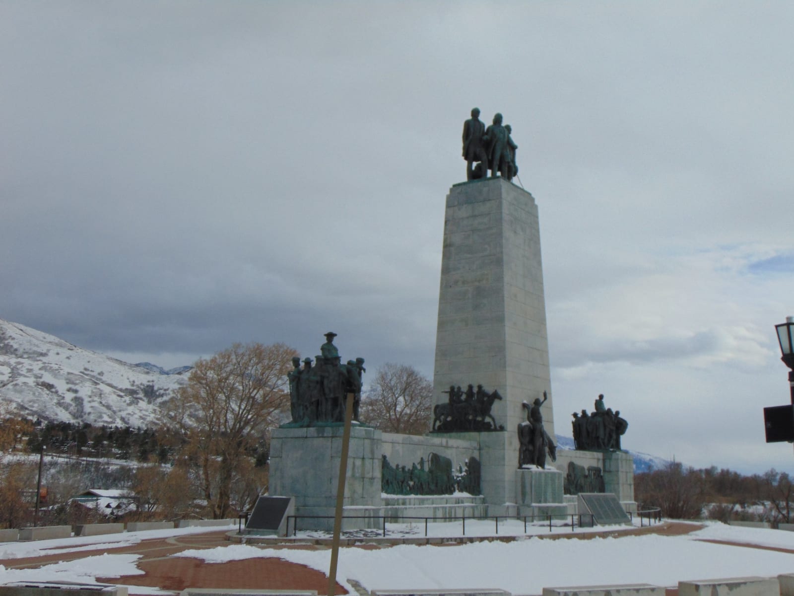 The This Is the Place Monument under a grey winter sky — a tall granite pylon topped by three standing bronze figures, with further bronze groups of pioneers, riders and ox teams set on the lower pedestals and relief panels worked around its base. Snow lies across the brick plaza and over the foothills behind, and the trees are bare.