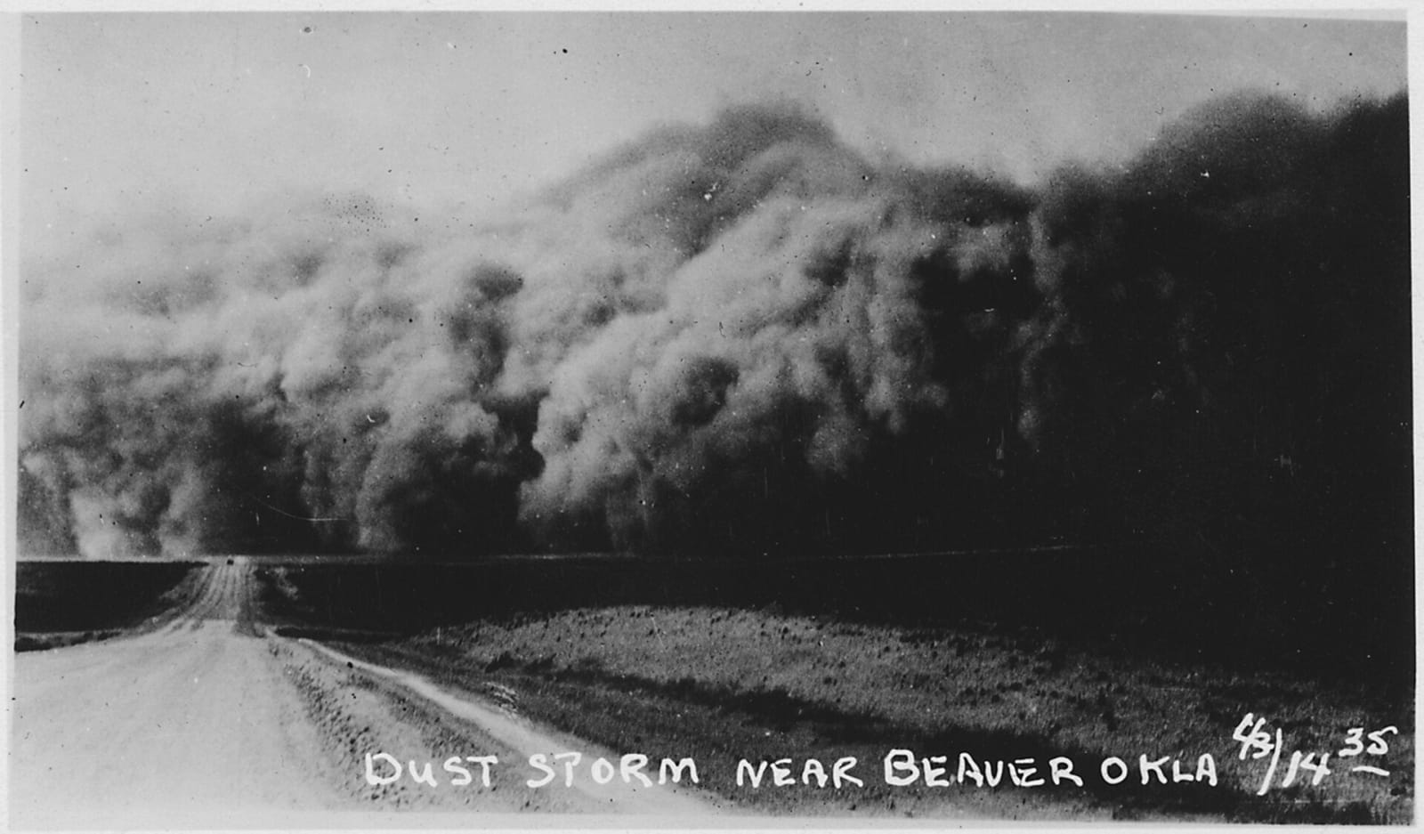 A black wall of dust rolling across the plains toward a dirt road near Beaver in the Oklahoma Panhandle in 1935, captioned Dust Storm Near Beaver Okla.