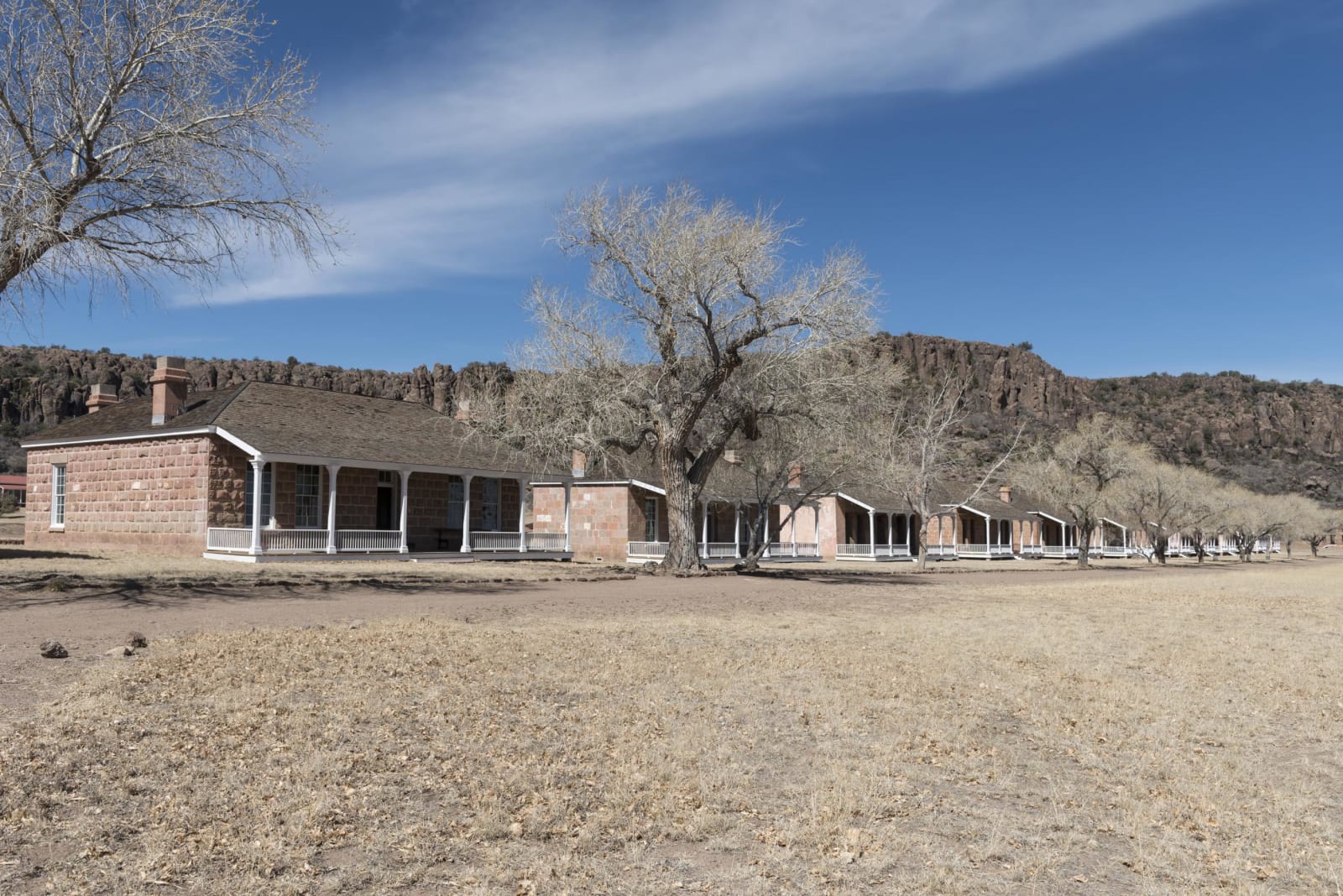 Restored stone barracks with long porches at Fort Davis National Historic Site, bare cottonwoods along the row and a rocky ridge rising behind.