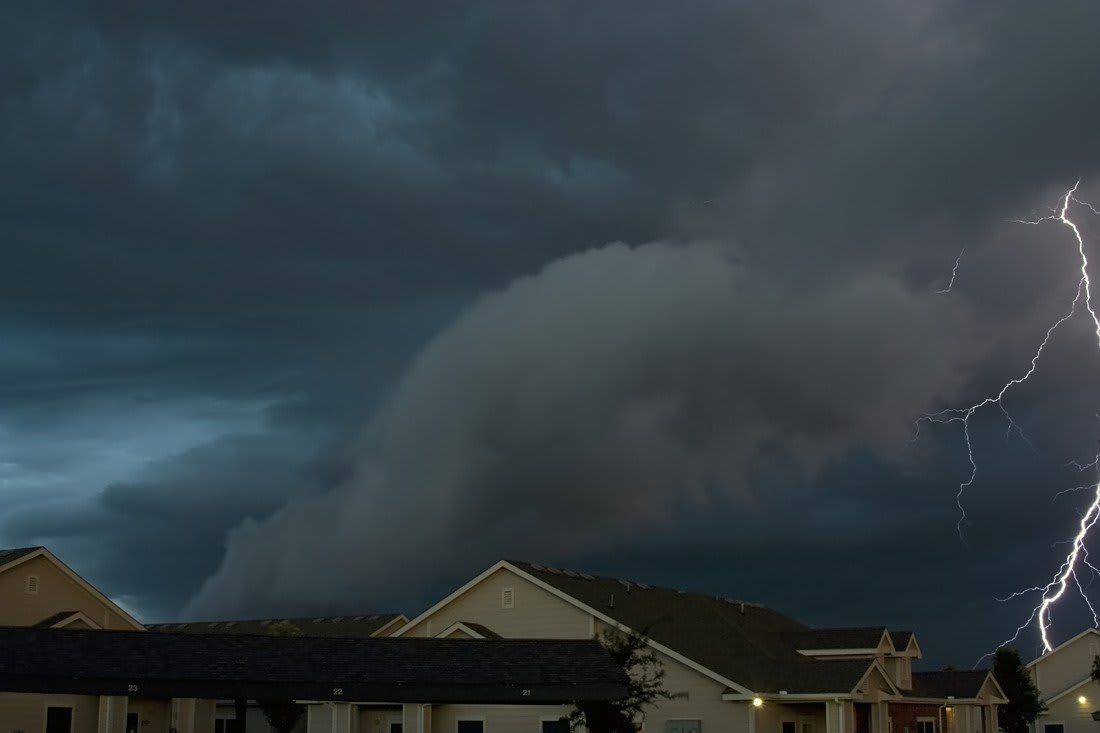 A towering shelf cloud rolling over rooftops in Lubbock at dusk, with lightning forking down at the right.
