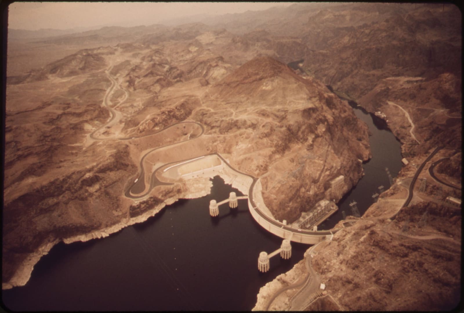 A May 1972 aerial view of Hoover Dam curving across Black Canyon, the waters of Lake Mead standing behind it at lower left with four intake towers rising from the reservoir, and the Colorado River continuing downstream to the right through bare desert mountains. A narrow pale band of exposed rock marks the shoreline where the water has dropped. The frame is a colour transparency scanned in its mount, its edges rounded and its colours shifted warm with age.