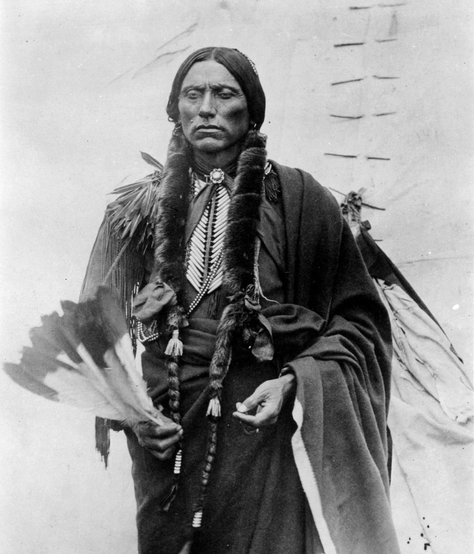 Quanah Parker standing in a studio portrait, wrapped in a long blanket with his hair in braids, in a black and white photograph.