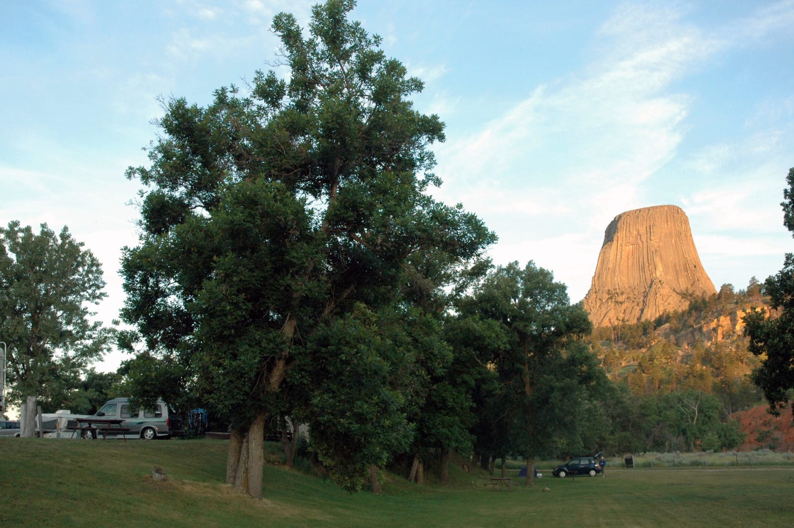 Devils Tower glowing orange at sunrise beyond the cottonwoods and parked campers of a campground at its foot