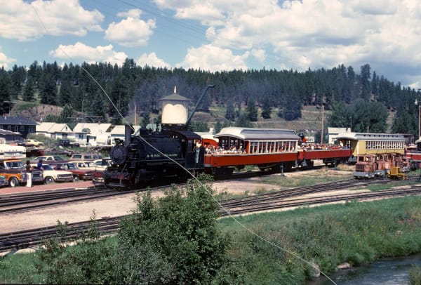 A steam locomotive pulling open-sided passenger cars past a water tower at the Hill City depot in 1978