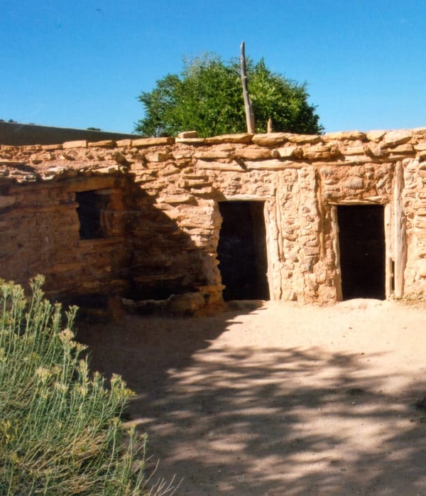 The reconstructed Ancestral Puebloan dwelling at Anasazi State Park Museum in Boulder, Utah — coursed sandstone walls with two dark doorways and a projecting timber lintel, sage in the foreground and a bare pole standing above the roofline.