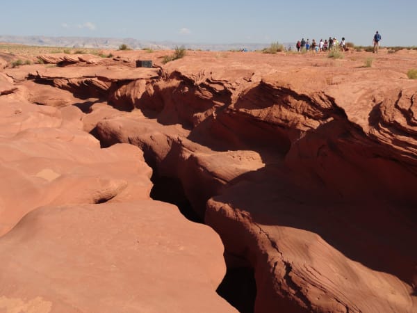 The slickrock bench above Antelope Canyon on the Navajo Nation east of Page, with the slot showing only as a narrow dark fissure winding away across the frame through cross-bedded sandstone worn into overhanging lips and fins. A line of visitors walks the rim toward the entrance in the distance and one hiker crosses the flat at the right, dry grass and low scrub run along the far edge, and the mesas above Lake Powell close a hazy horizon. The carved corridor and the shaft of light the canyon is known for are below the surface and not visible in the frame.
