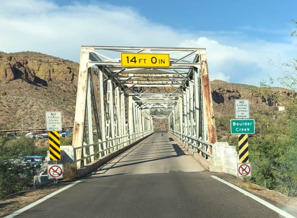 A steel through-truss bridge on the Apache Trail seen head-on from the middle of the roadway, its white-painted portal carrying a yellow clearance sign lettered 14 FT 0 IN, the trusses running away over the deck to a vanishing point. Square signs at both approaches read NO FISHING DIVING LOITERING ON BRIDGE, a green sign at the right names Boulder Creek, and black-and-yellow chevron markers stand on white concrete abutments at each side. Layered volcanic hills carrying palo verde and saguaro rise behind on both banks under a blue sky with cumulus. The narrow unpaved cliff-edge section the road is known for is not in the frame.