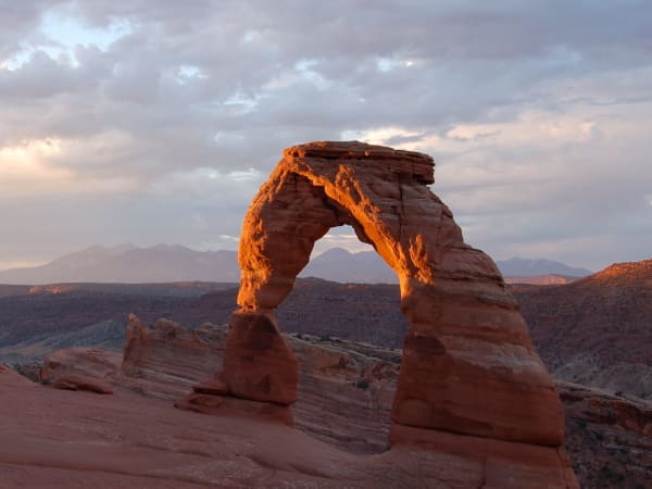 Delicate Arch standing free on a slickrock bench, lit deep orange by low sun, its two thick legs tapering to a curved span. Snow-streaked peaks stand on the horizon behind, and heavy grey cloud fills the upper half of the frame.