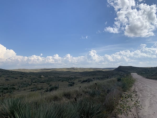 Yucca-dotted ridges and canyons of the Arikaree Breaks fall away beside a dirt road under a cloud-flecked sky.