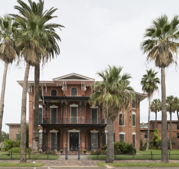 The red brick Ashton Villa on Broadway in Galveston, with iron galleries on two floors, framed by palms behind an iron fence.