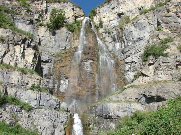 A tall waterfall fanning down a grey limestone cliff — the water splitting into several strands over a rust-stained face at centre, ledges of pale banded rock stepping up on both sides with shrubs and a lone conifer rooted in them, and a small group of hikers standing on the rock apron at the right.