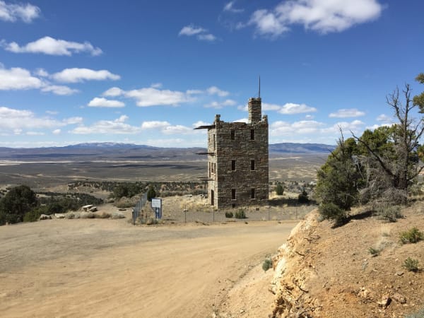 Stokes Castle on its bluff above Austin — a three-storey tower of rough hand-hewn granite with a battlemented top, empty window openings and the stubs of its balcony rails still projecting from the walls, standing inside a chain-link fence at the edge of a gravel turnaround. Beyond it the Reese River Valley runs pale and open to a long range on the horizon, with a scatter of low buildings and utility poles at the valley edge below. The town's main street is not in the frame.