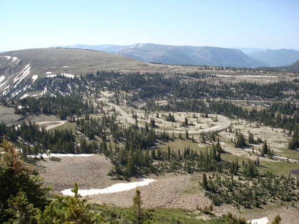 A high alpine basin seen from above the treeline, a paved road switchbacking through scattered spruce and open rock meadow with patches of old snow. A long flat-topped ridge runs across the middle distance and hazy ranges recede beyond under a pale sky.