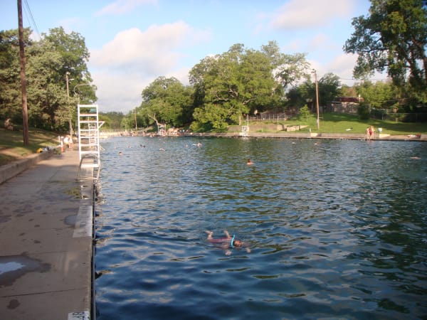 The long clear pool at Barton Springs in Austin, with a few swimmers, a diving board and trees along the far bank.