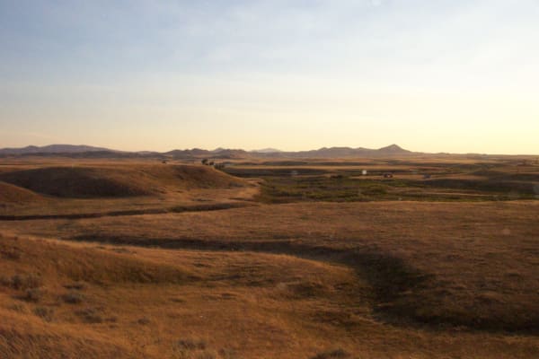 The shallow grassy bowl of the Bear Paw battlefield in low evening light, dry prairie rolling to distant buttes