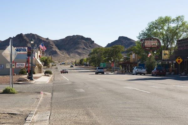 Beatty's main street, with US-95 running straight through town between low storefronts under bare rocky hills. On the left stand the Exchange Club Motel, a hardware store and a lamp standard with flags; on the right an oval Exchange Club Casino-Motel neon, a Sourdough Saloon sign, awnings and parked pickups. A yellow diamond sign warning of burros stands at the right kerb, one car comes up the highway, and empty asphalt fills the foreground.