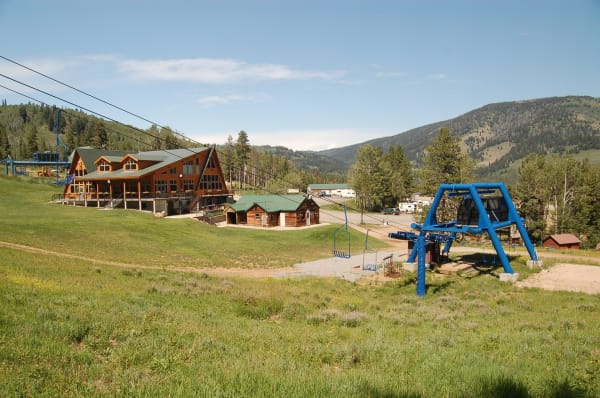 A ski area base in summer with no snow on the ground, a timber lodge with a green roof and a wide deck standing above a mown green slope, a smaller log building beside it, and a blue chairlift terminal at the right with empty chairs parked on the line. Forested ridges rise behind.