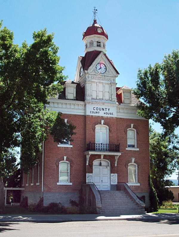 A red brick county courthouse photographed head-on from the empty street, its white stone trim weathered and peeling, a clock tower rising to a dark red dome and weathervane, COUNTY COURT HOUSE lettered across the frieze, and two curving flights of steps climbing to arched double doors between tall shade trees.