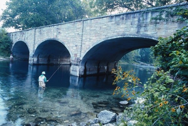 An angler wading clear spring water beneath the arched stone bridge at Bennett Spring State Park.