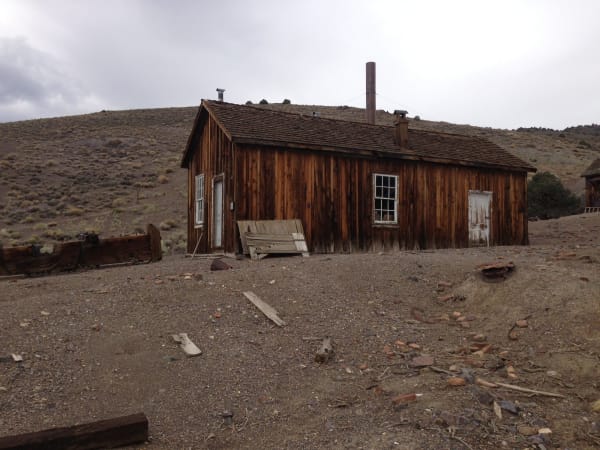 A weathered board-and-batten cabin at Berlin, its unpainted boards silvered and streaked rust-brown, with a shingled roof carrying two stovepipes, two white-framed windows and two plank doors. Loose boards lean against the wall, cut timber and broken firebrick lie scattered across the bare gravel in front, a low timber footing runs along the left edge, and a sagebrush hillside rises behind under flat grey overcast. The ichthyosaur quarry and its fossils are not in the frame.