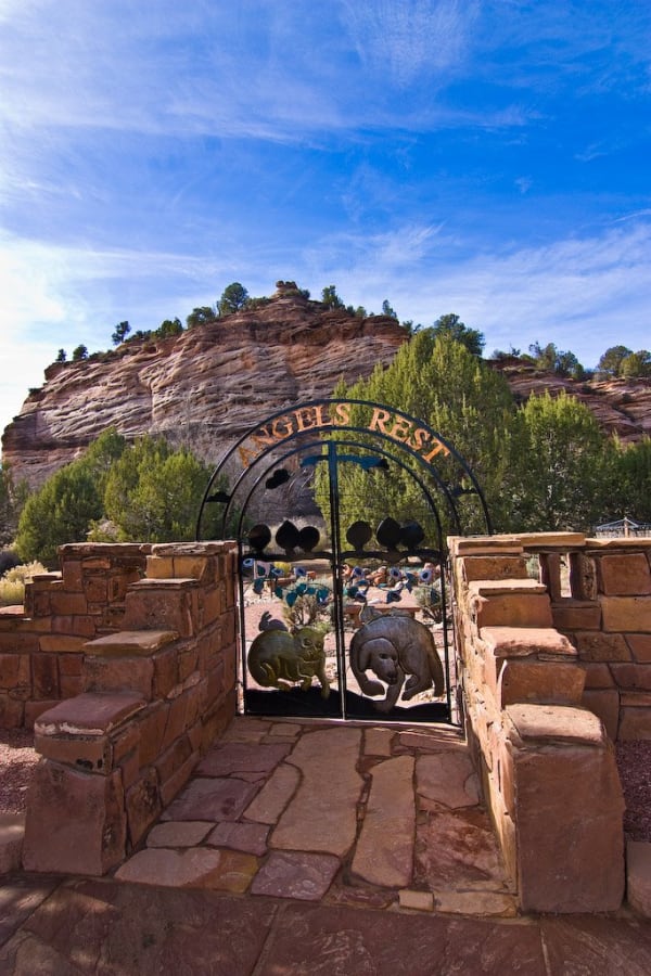 The gate of Angels Rest, the pet cemetery at Best Friends Animal Sanctuary — twin iron arches lettered ANGELS REST in copper, cut-metal paw prints along the tops of the gates and bird silhouettes hung from the inner arch, and a cat and a dog cut in sheet metal filling the two leaves. Flagstone paving runs up between low walls of stacked red sandstone. Beyond the gate stand metal flowers on stems, a timber bench and gravel ground; a banded sandstone cliff with junipers on its ledges rises behind. None of the sanctuary's animal areas are in the frame.