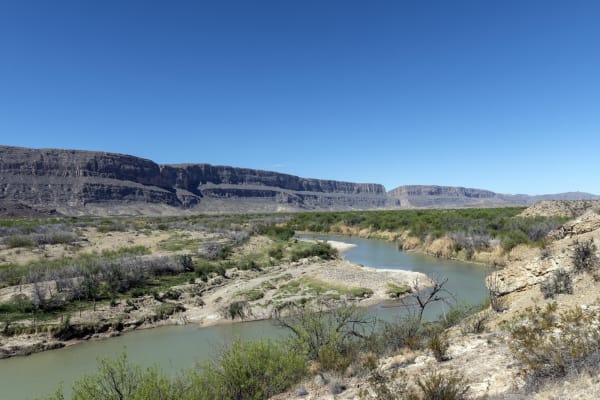 The Rio Grande curving through green brush below pale limestone bluffs in Big Bend National Park, under a clear blue sky.