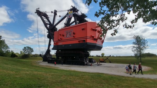 Big Brutus, a giant orange-and-black electric shovel with a tall boom, towering over visitors on a gravel path at West Mineral.
