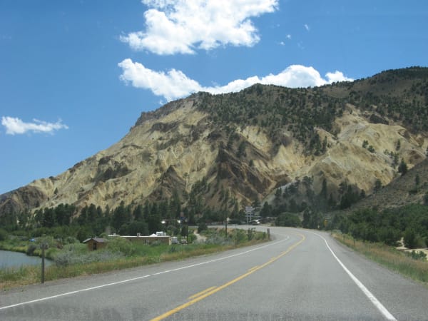 A two-lane highway seen from the road itself, curving right beneath a steep hillside of pale yellow and ochre rock cut into ribs and gullies. Pines and a river line the left side, a few small roadside buildings sit at the foot of the slope, and cumulus stands over the ridge.