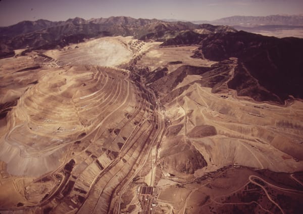 A July 1972 aerial view of the Bingham Canyon open-pit mine, concentric benches stepping down the left half of the frame, waste-rock dumps and switchbacked haul roads across the right, mill buildings on the valley floor below, and range after range receding into haze on the horizon.