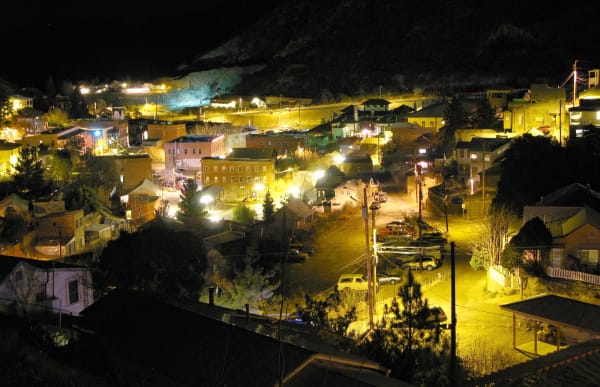 Bisbee at night from the slope above — the town filling the canyon floor and climbing both walls, brick commercial blocks lit orange by sodium lamps in the middle distance, frame houses and sheds stepped up the hillsides on either side, and pickups parked along the streets below. Power poles and slack wire cross the foreground, and a bare pale slope stands out cold and blue under floodlight above a lit yard at the top of the frame. The Copper Queen works and the open pits are not in the frame.