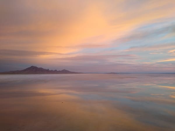 The Bonneville Salt Flats under a thin sheet of standing water at sunset, the flooded surface mirroring pink and orange cloud almost exactly, with a low dark range along the horizon at left.