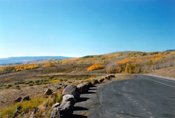 Highway 12 curving right along a boulder-lined pullout on Boulder Mountain, Utah — aspen in full autumn gold across the slopes and blue ridges receding into the distance.
