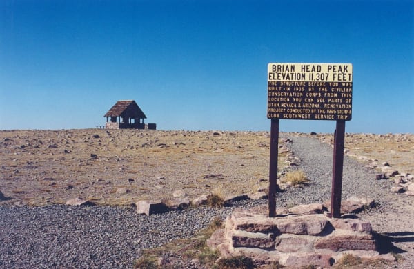 The summit of Brian Head Peak — bare, stony alpine ground running flat to the horizon under a deep blue sky, with the open stone-and-timber shelter the Civilian Conservation Corps built in 1935 standing on the skyline at left. At right a yellow interpretive sign on steel posts, set in a flagstone base, reads BRIAN HEAD PEAK, ELEVATION 11,307 FEET, and records the 1935 CCC construction and a 1995 Sierra Club renovation. A gravel track runs past it toward the shelter. The town of Brian Head itself is not in the frame.