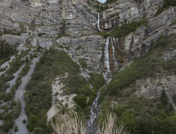 Bridal Veil Falls in Provo Canyon — the upper cascade dropping through a notch in the grey limestone cliff and the lower fall spreading in white strands down a broad rust-stained face below it, the stream running out over boulders into the trees at the bottom of the frame. A scree gully and brush-covered slopes fill the left half under flat grey light.