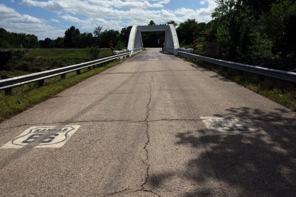 Old Route 66 pavement, painted with route shields, leading to the white concrete arch of the Brush Creek Bridge.