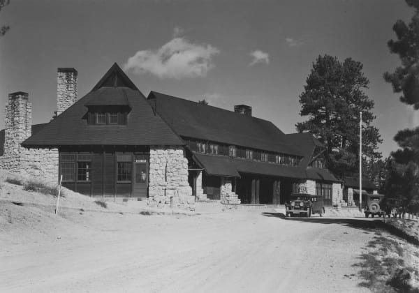 An archival black-and-white photograph of Bryce Canyon Lodge — steep shingled roofs and gables above rough-cut stone walls and chimneys, a dormer over the near wing, and a small sign reading BARBER SHOP beside one door, with a covered entrance porch on log posts further along the facade. Period touring cars stand at the entrance and ponderosa pines rise at right, above a broad unpaved forecourt.