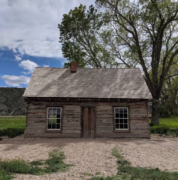 A small single-story cabin of weathered hewn logs photographed straight on, with a wood shingle roof, a red brick chimney, a plank door at the center and a white framed sash window on each side. A large cottonwood rises behind it and open pasture runs back to a hillside.